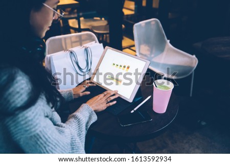 Crop young beautiful female freelancer in glasses and in headphones watching statistics on screen of tablet working at table at cafe