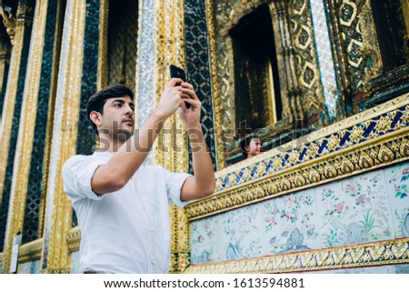 Serious spanish male travel blogger standing near golden temple making picture of asian notable places and architecture, man tourist shooting video for blog using smartphone camera outdoors