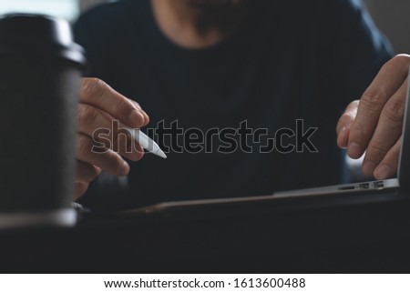 Casual man, freelance with stylus pen writing on digital tablet screen, working on laptop computer with cup of coffee on office desk. Graphic or web designer working his web design project, dark tone