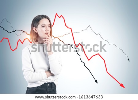 Thoughtful sad young woman in casual clothes standing near gray wall with falling graphs. Concept of financial crisis and loss