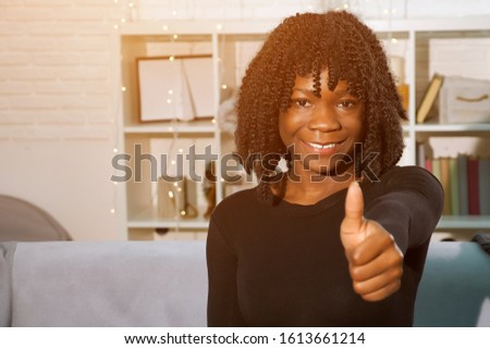 beautiful young african woman in black pullover raises hand and shows thumb-up sitting on grey sofa at home, sunlight