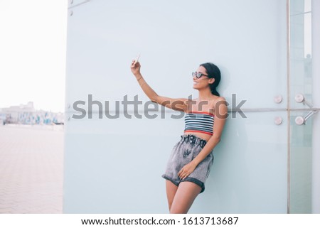 Happy female Generation Y in trendy sunglasses smiling while posing for shooting influence vlog and share to medial social networks, cheerful hipster girl enjoying online video translation on cellular