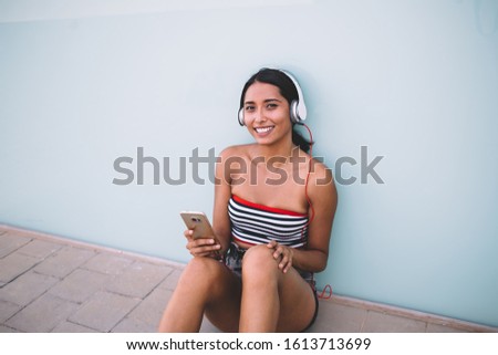 Portrait of happy millennial woman holding smartphone technology and laughing during leisure for listening podcast from popular medial website, pretty Spanish hipster girl in electronic headphones