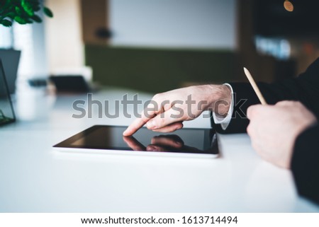 Crop side view of young male office worker in suit browsing on tablet while sitting at desk at urban office in daylight