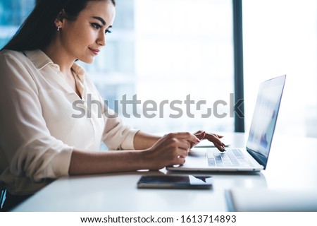 Side view of beautiful young woman smiling and browsing modern laptop while sitting at table against window and working in office
