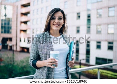 Half length portrait of beautiful young caucasian businesswoman in spectacles having work break outdoors, smiling female student holding books and coffee to go cup rest on urban setting background