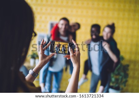 Anonymous woman using smartphone to take pictures of happy multiracial friends having fun together against bright wall in stylish apartment