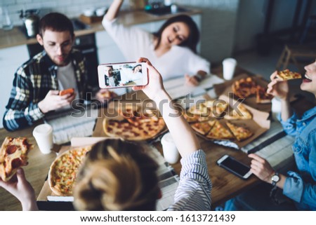 High angle selective focus of unrecognizable female taking picture of cheerful mates having fun at table while eating pizza during friends meeting at home