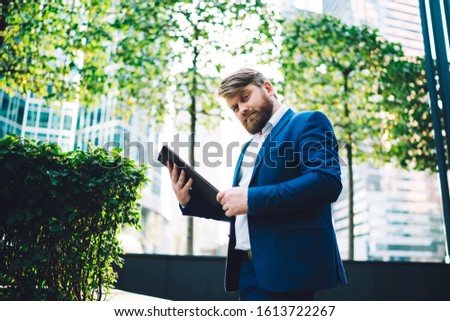From below of confident modern bearded businessman in formal blue suit using tablet while standing on street with green trees and office buildings in background 