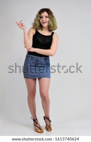 A horizontal photo concept of a young woman in a black t-shirt and blue skirt is standing in front of the camera on a white background. Full-length portrait of a pretty girl with beautiful curly hair.