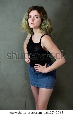 Concept vertical photo of a young woman in a black T-shirt and blue skirt standing straight. Portrait of a pretty student girl with beautiful curly hair in front of the camera on a gray background.