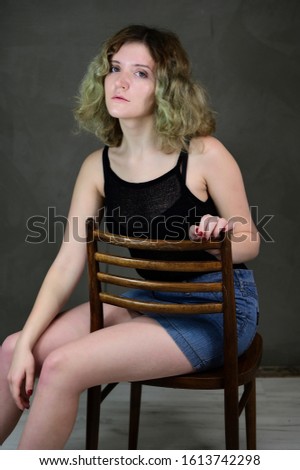 Portrait of a pretty student girl with beautiful curly hair in front of the camera on a gray background. Concept vertical photo of a young woman in a black T-shirt and blue skirt sitting on a chair.