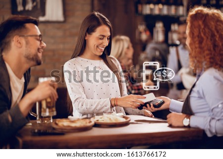 Young happy woman using smart phone while paying for a bill to a waitress in a bar. 