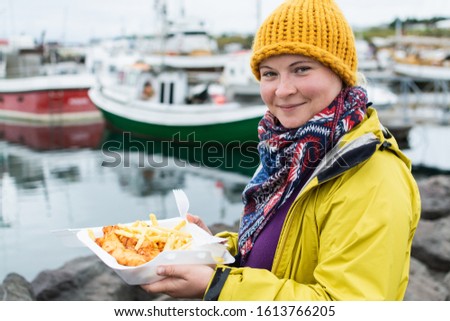 Young woman in yellow raincoat eats a portion of fish and chips in the port of Husavik, Iceland. Stock photo.