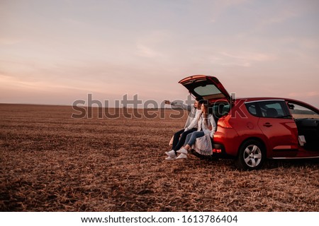 Young Happy Couple Dressed Alike in White Shirt and Jeans Sitting at Their New Car Trunk, Beautiful Sunset on the Field, Vacation and Travel Concept Royalty-Free Stock Photo #1613786404