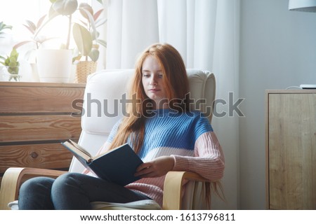 portrait of pretty young girl with red hair sitting in comfy chair reading a book Royalty-Free Stock Photo #1613796811