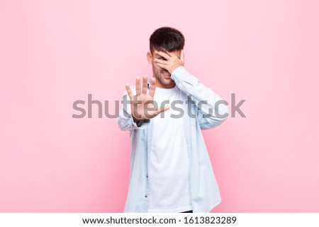 young handsome man covering face with hand and putting other hand up front to stop camera, refusing photos or pictures against pink wall