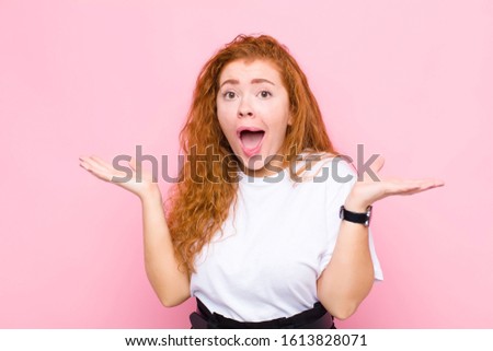 young red head woman looking shocked and astonished, with jaw dropped in surprise when realizing something unbelievable against pink wall