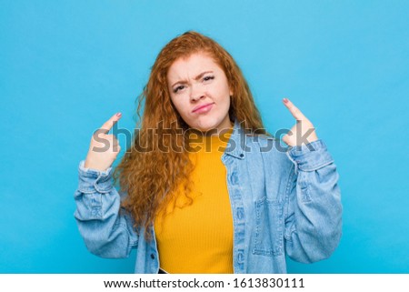 young red head woman with a bad attitude looking proud and aggressive, pointing upwards or making fun sign with hands against blue wall