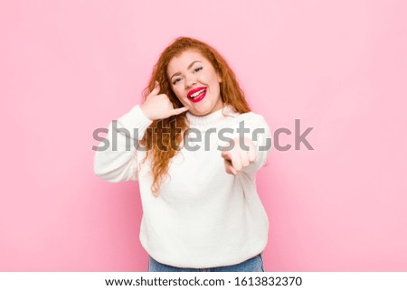 young red head woman smiling cheerfully and pointing to camera while making a call you later gesture, talking on phone against pink wall