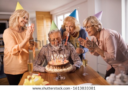 Group of happy senior friends celebrating Birthday at home. Man is blowing candles on a cake. 