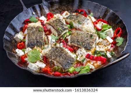 Traditional fried Greek cod fish filet casserole with feta cheese and vegetable as closeup in a rustic wrought iron pan