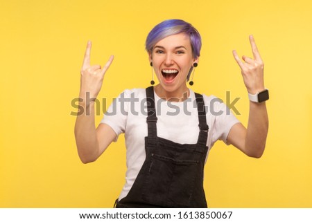 Portrait of delighted funky hipster woman with violet short hair in overalls showing rock and roll gesture, crazy punk sign, devil horns and shouting with happiness. yellow background, studio shot