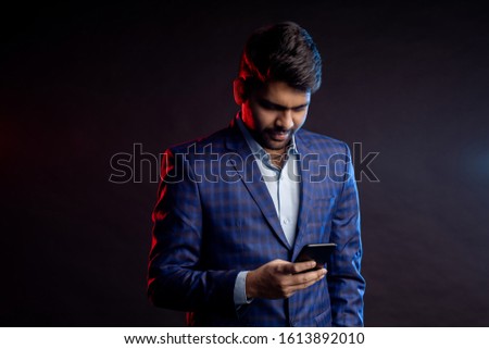 Portrait of confident stylish young indian man in formalwear, holding mobile phone, looking at screen, standing against black background. Successful businessman. Business, technology.