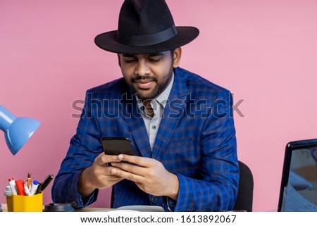 Indian businessman wearing suit, black hat, looking at screen of mobile phone, waiting for important call, reading news, sending email, sitting at working desk in office, over pink background.
