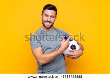 Man over isolated yellow background with soccer ball and pointing to the lateral