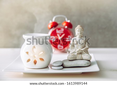 How to find balance between busy life and healthy mental health. Taking a break to sit and meditate concept. Buddha figure with aroma lamp and clock on the background.