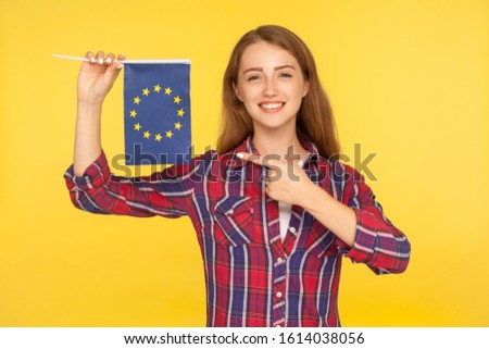 European Union. Portrait of positive ginger girl in checkered shirt pointing at European flag, citizen of EU smiling at camera, showing patriotic banner. studio shot isolated on yellow background