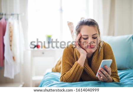 Bored young woman with smartphone lying on bed indoors at home.