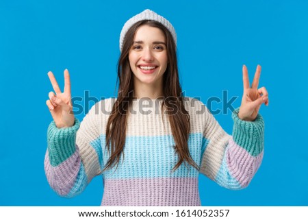 Happy and carefree cute, charismatic brunette female in winter sweater, hat, showing two peace signs and smiling broadly, photographing on winter holidays, mountain ski luxury resort, blue background