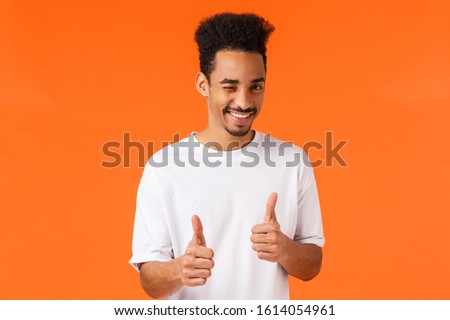 You can do it, say yes. Cheerful and confident young supportive african-american man in white t-shirt, wink and smile, showing thumbs-up gesture, rooting, encourage everything good, nice work