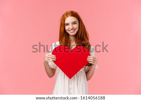 Tenderness, love and romance concept. Romantic charming redhead girlfriend prepared cute gift valentines day, showing red heart card and smiling, express affection and sympathy, pink background