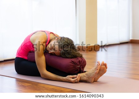 Middle aged woman on pink tank top in paschimottanasana pose resting chest on bolster at bright studio. Female yogi on seated forward bend asana. Therapeutic exercise concept Royalty-Free Stock Photo #1614090343
