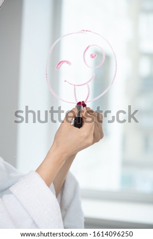 Hand of young female in white bathrobe with crimson lipstick drawing grimacing funny face on mirror