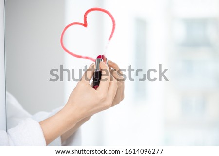 Hand of flirty female in white bathrobe drawing heart with crimson lipstick on mirror in the bathroom