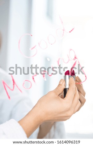 Hand of young woman holding lipstick of crimson color close to mirror while writing good morning