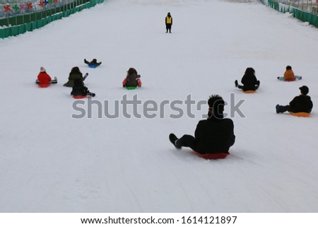 People are sledding down on the snow slide. And security guards in yellow are watching.