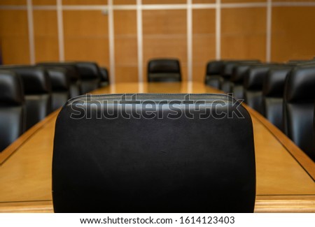 Close up of the back of a large black chair in conference room with other chairs facing toward it. The light brown conference table visible.  In the background is a  a metal and wood grid pattern.  Royalty-Free Stock Photo #1614123403