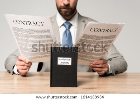 cropped view of businessman reading contracts near book with intellectual property title isolated on grey