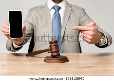 cropped view of lawyer pointing with finger at smartphone with blank screen, and gavel on desk isolated on grey