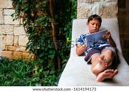 Cute little boy using tablet in sunlit park. Child with modern device playing computer games. Kid with contemporary gadget surfing web, watching movies. Modern recreation, childhood activity