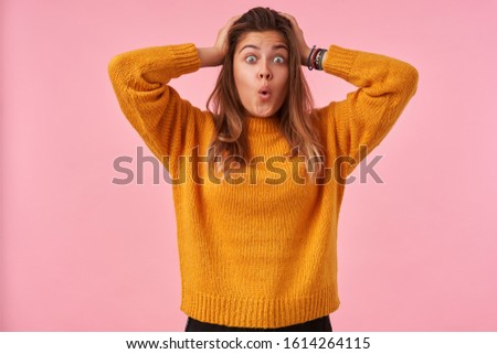 Shocked young brunette female with natural makeup clutching her head with raised hands while looking amazedly at camera with wide eyes opened, isolated over pink background