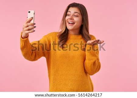 Positive young lovely brown haired woman with natural makeup raising hand with mobile phone while making selfie, smiling pleasantly with raised palm while posing over pink background