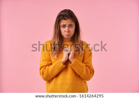Upset young attractive brunette female with casual hairstyle folding her raised hands and looking pitifully at camera with pouted lips, posing over pink background