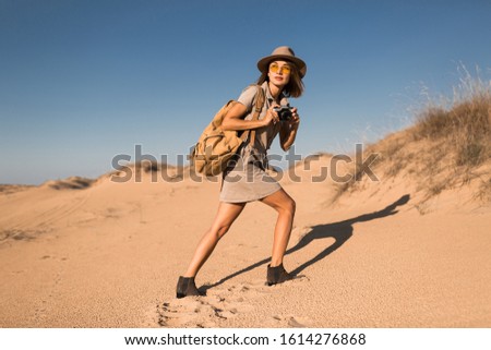 stylish young woman in khaki dress walking in desert sand, traveling in Africa on safari, wearing hat and backpack, taking photo on vintage camera, exploring nature, hot summer day, sunny weather