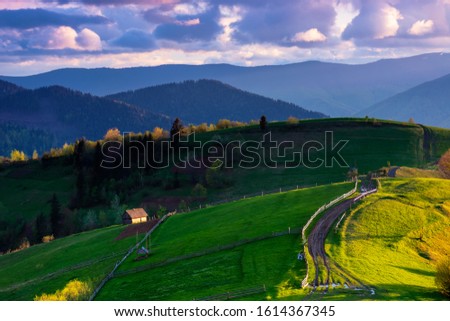 mountainous rural landscape in evening light. wooden fence along the path through rolling hills in fresh green grass. beautiful scenery in springtime. purple clouds on the sky